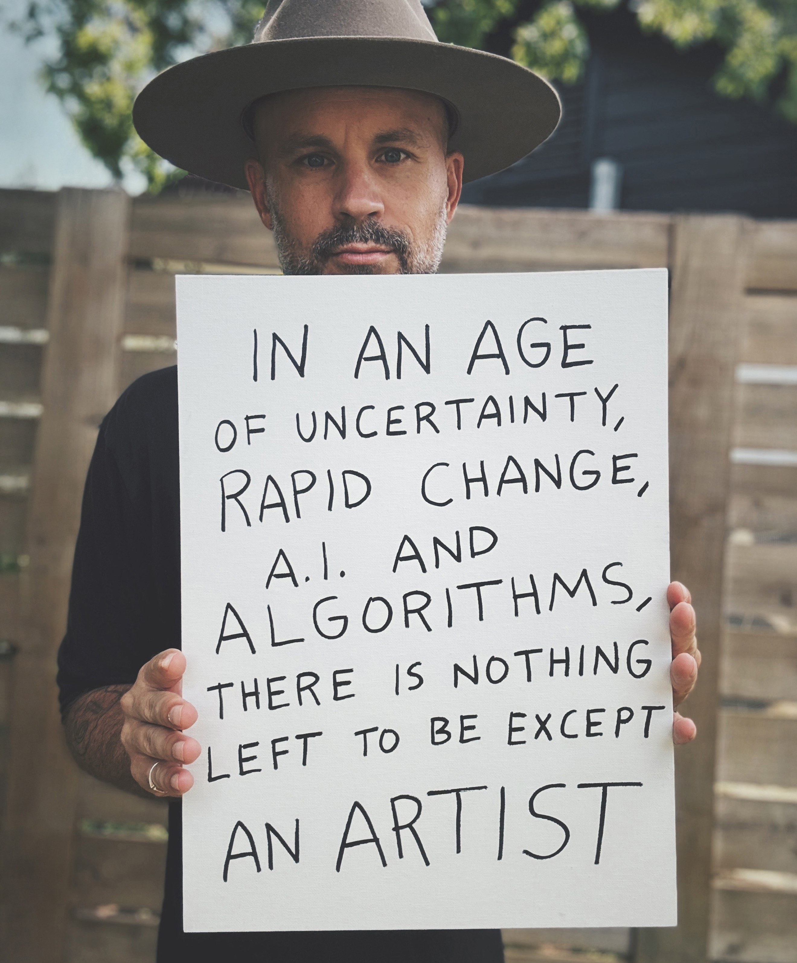 James McCrae holding a sign: In an age of uncertainty, rapid change, AI and algorithms, there is nothing left to be except an artist